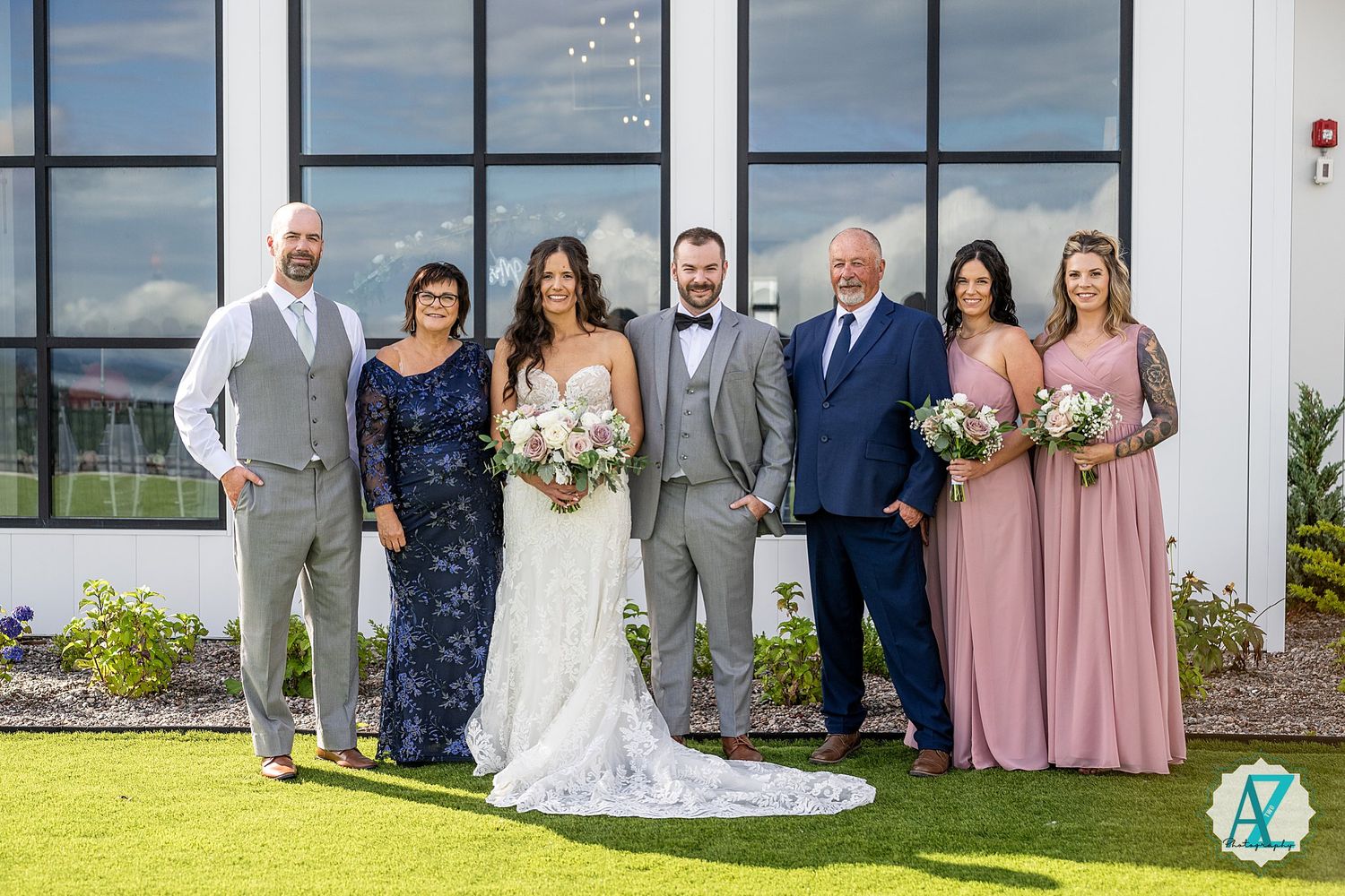 Elegant wedding party gathers on lawn for formal group photo against modern architectural backdrop.