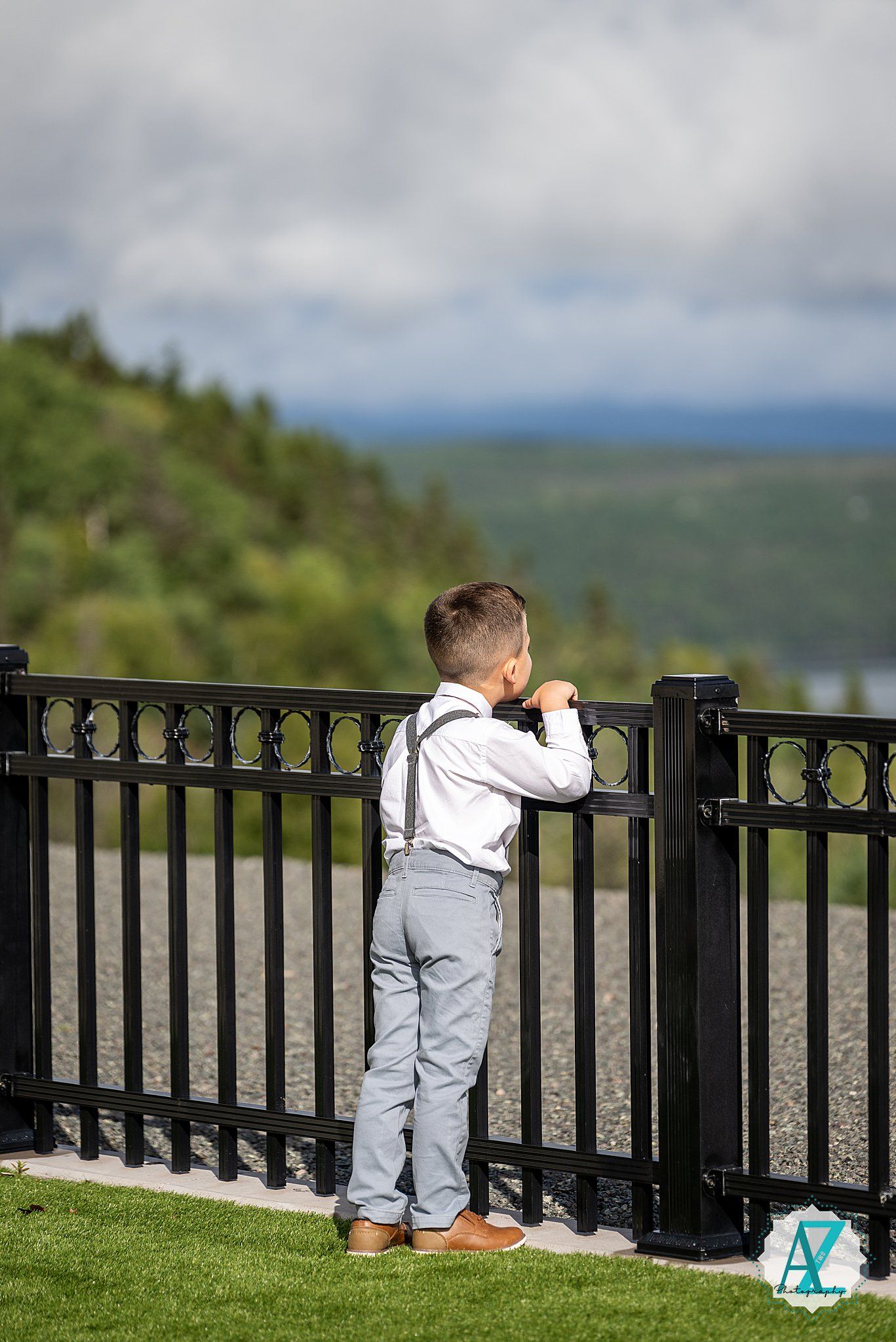 Children in gray suits sit along a black railing overlooking a scenic mountain landscape view.