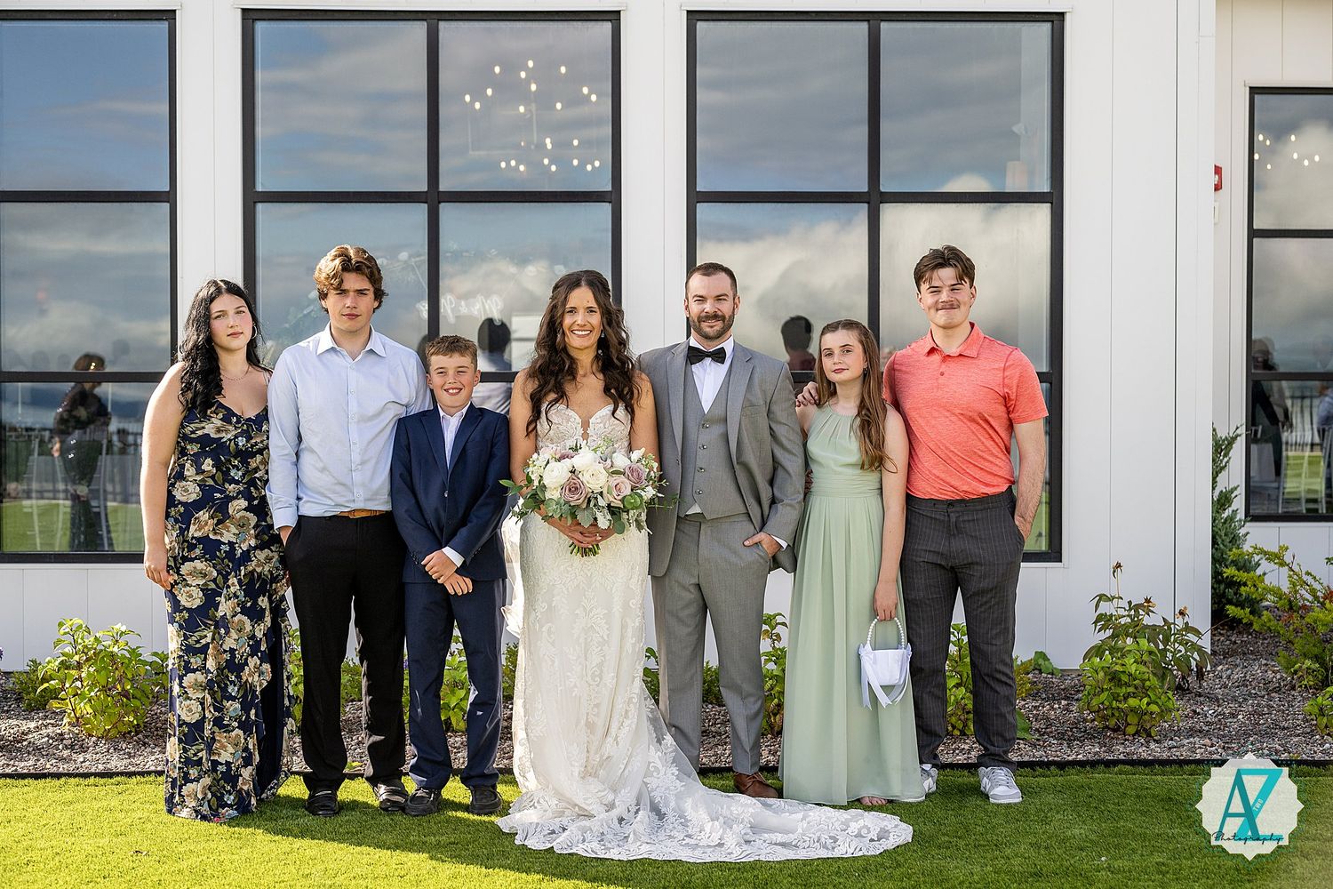 Wedding party members lined up outdoors against a white building with large windows for a group photo.