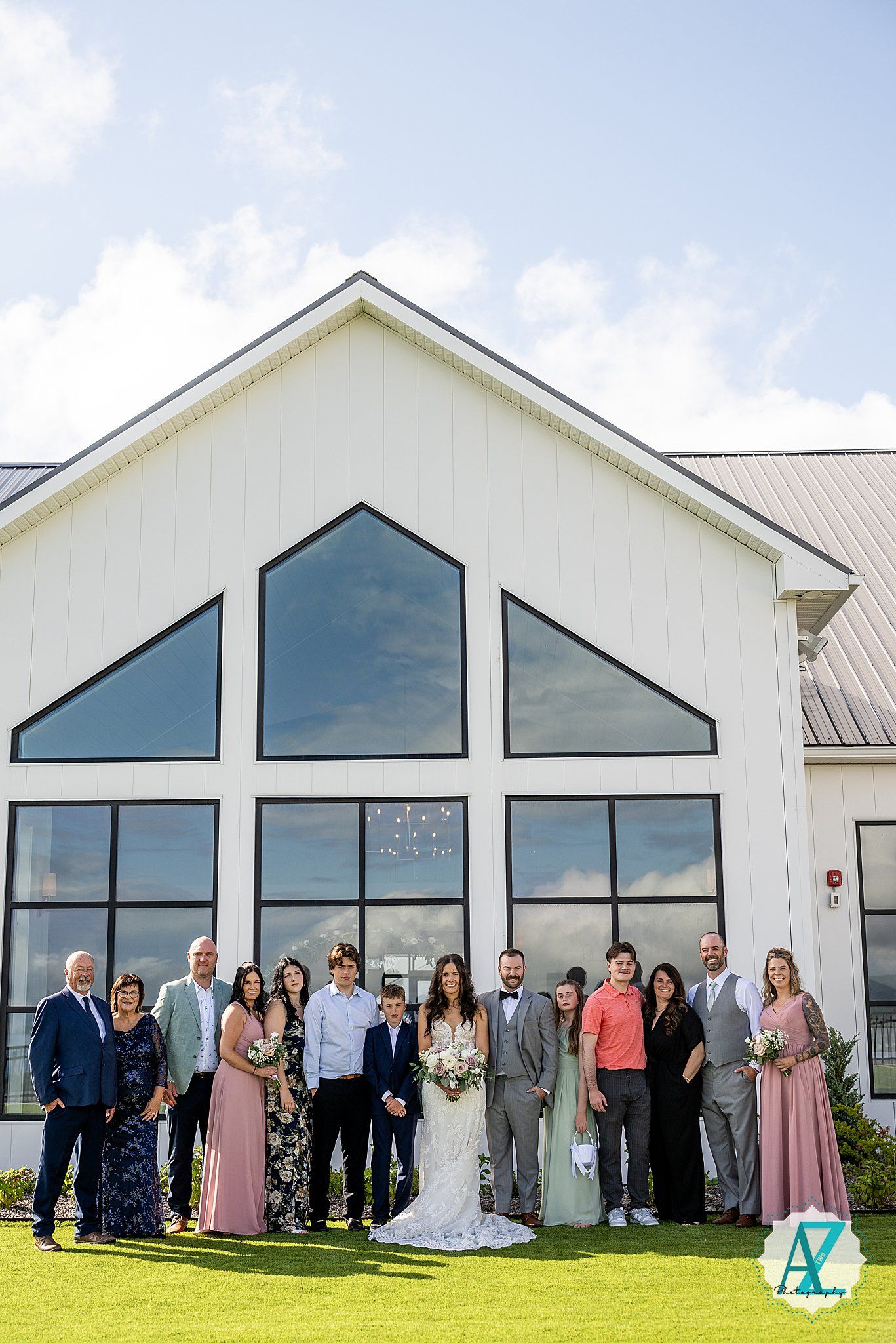 Wedding party lines up against modern white building facade with dark windows under blue sky on grassy lawn.