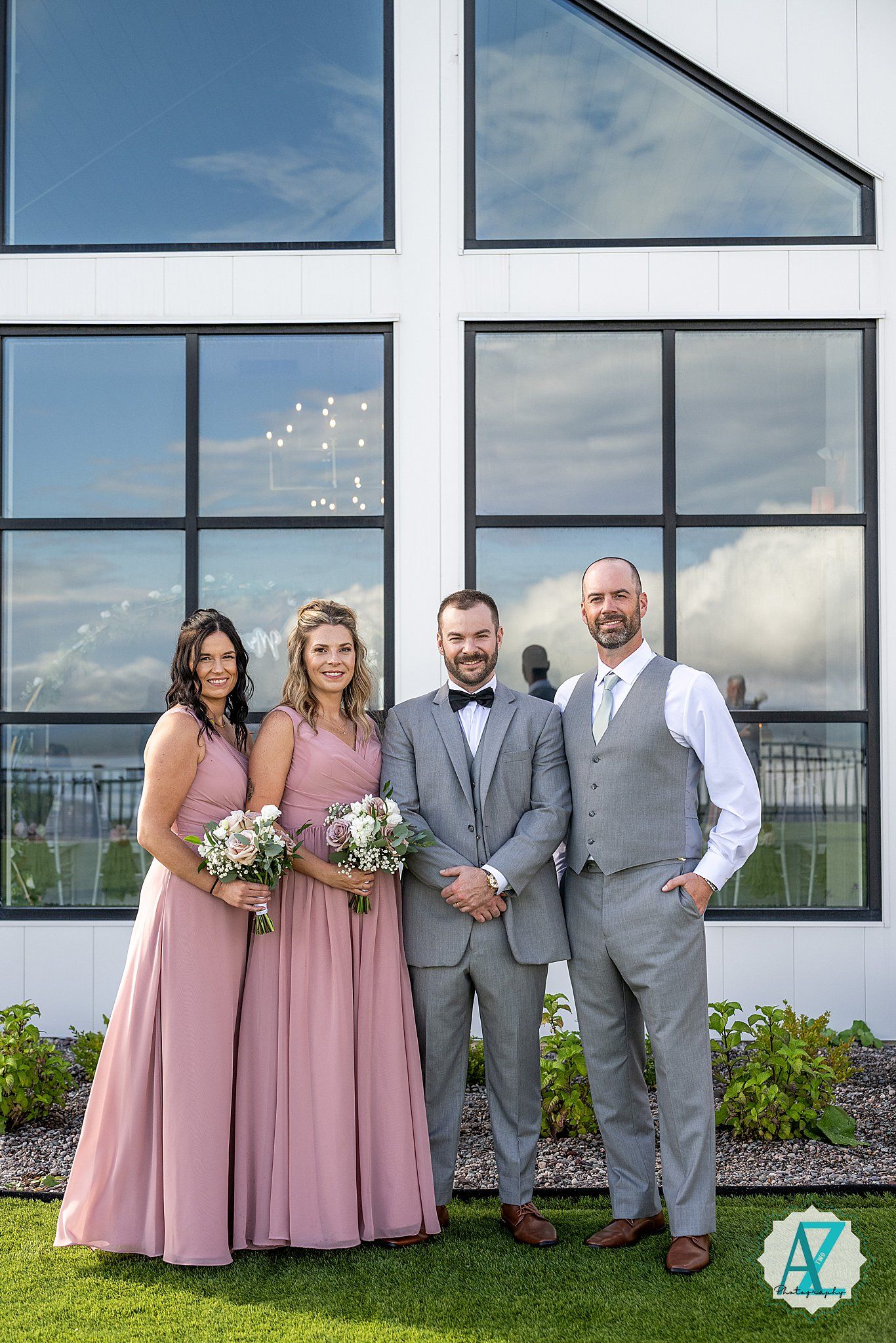 A wedding party in pink dresses and grey suits poses outdoors against a modern glass building backdrop on a sunny day.