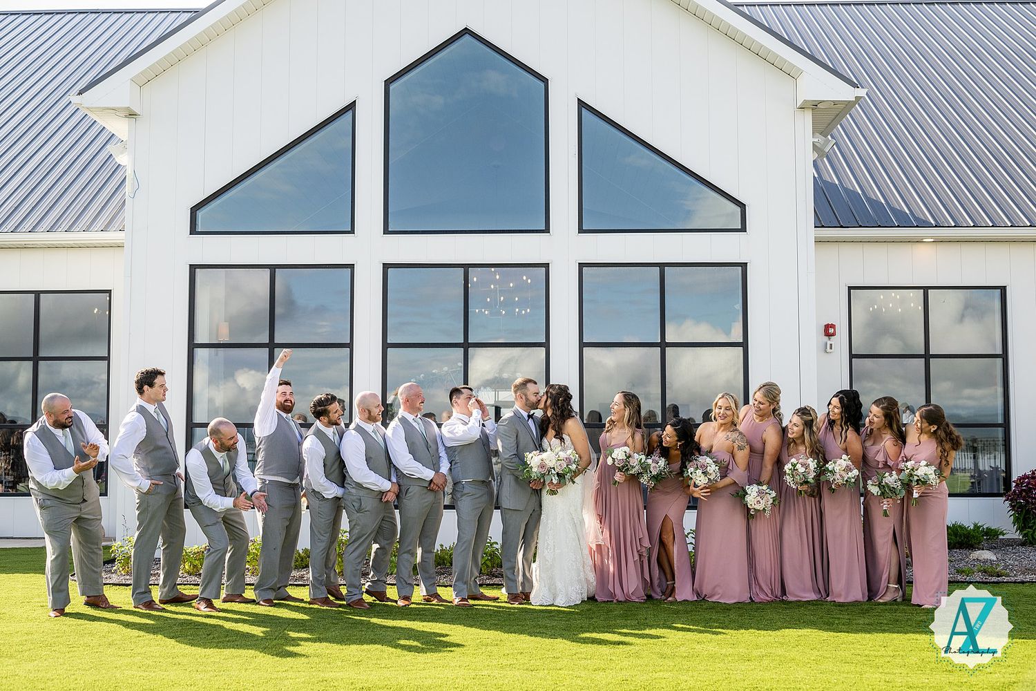 Large wedding party lined up in pink and grey attire against a white modern building with glass windows.