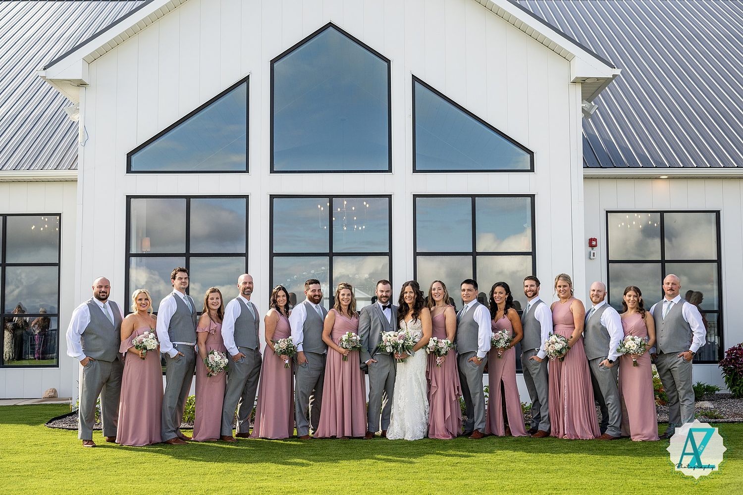 Wedding party wearing pink and grey attire poses in line against white church building with large windows.