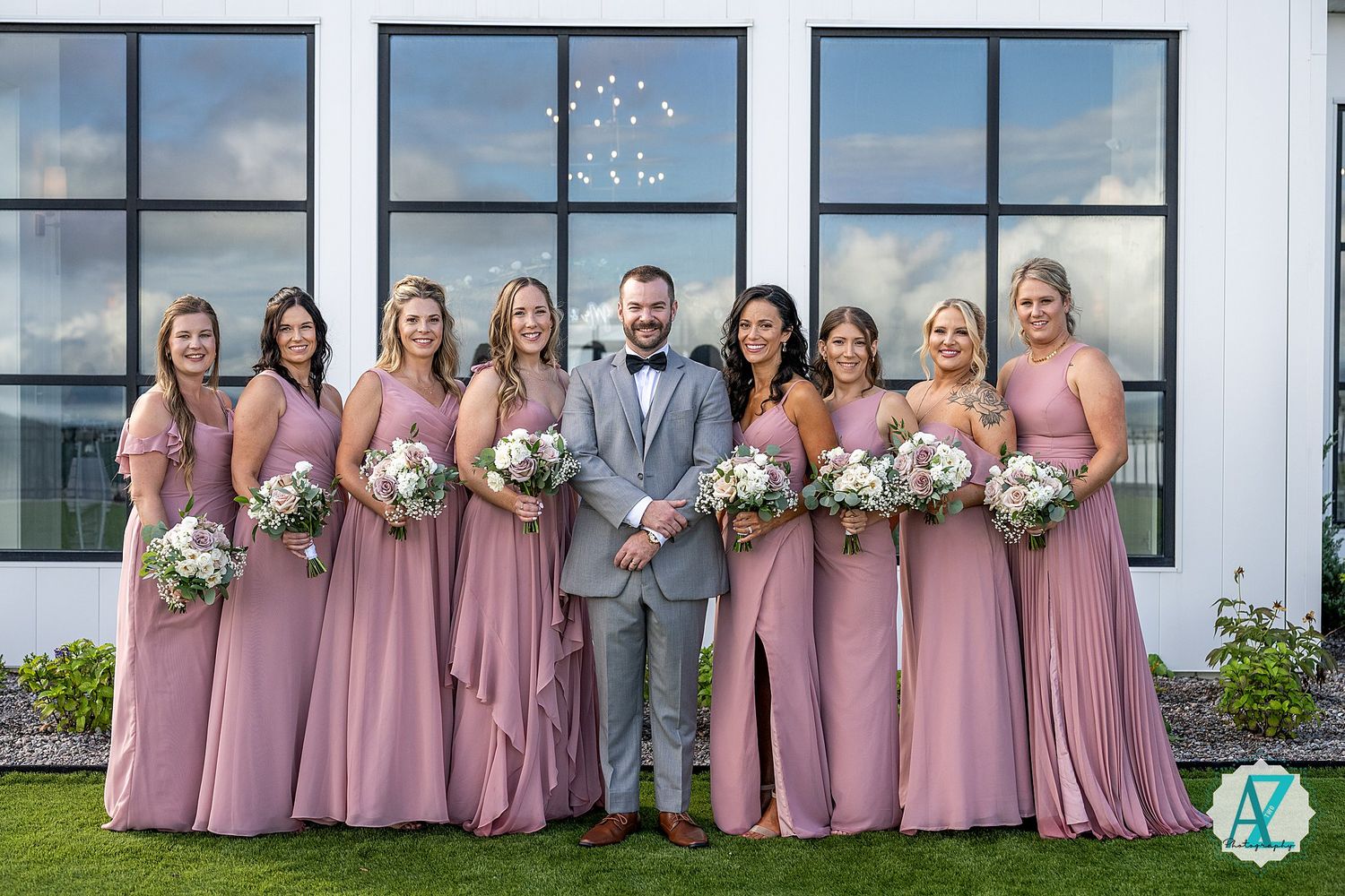Wedding party in pink dresses and grey suits poses in front of a white building with windows.