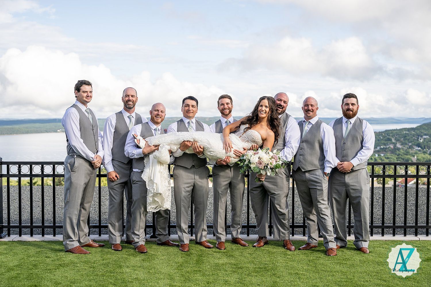 Wedding party in gray suits and dresses sits casually along black railing with mountain backdrop.