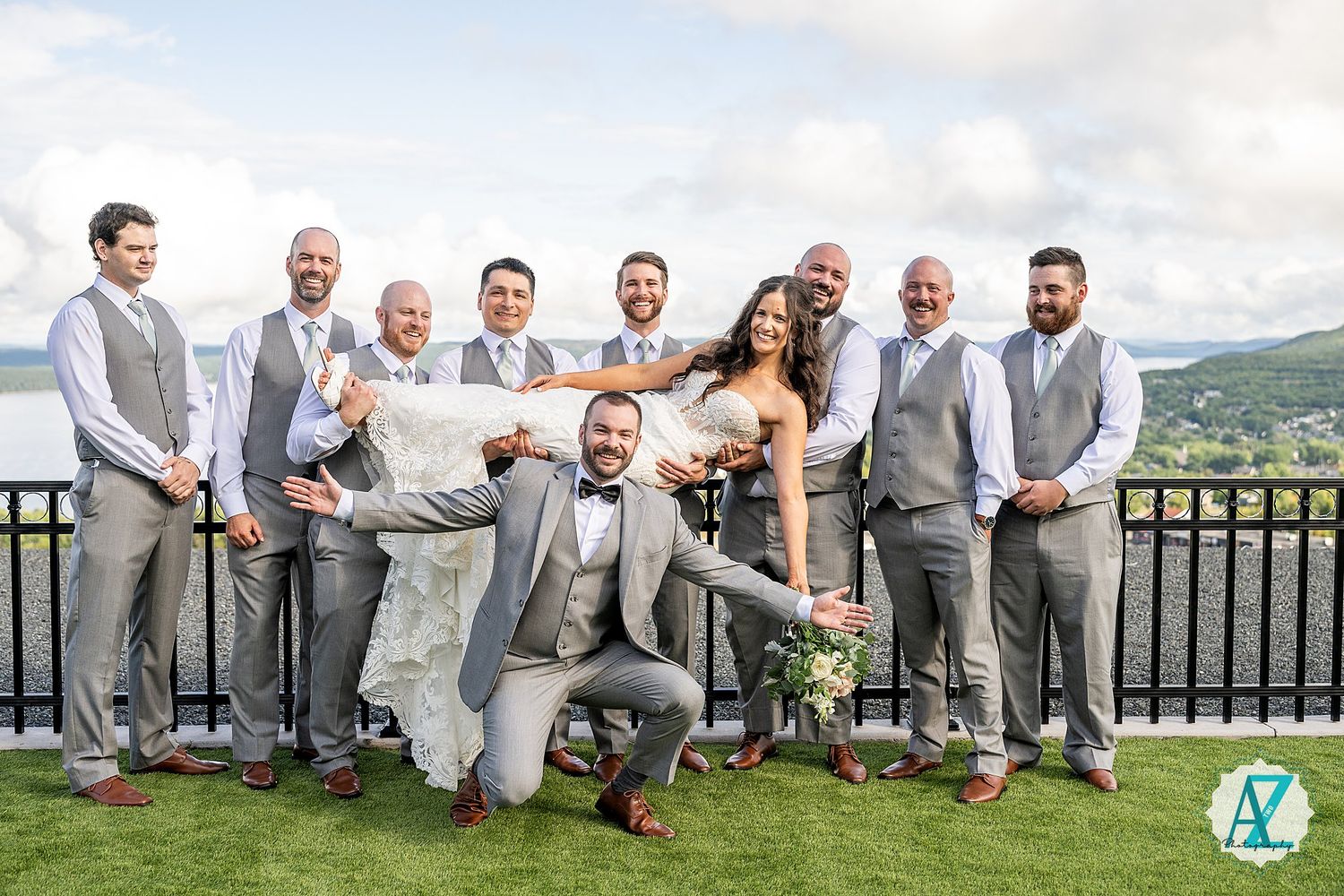 Wedding party in gray suits and formal attire pose together on balcony overlooking mountain landscape.