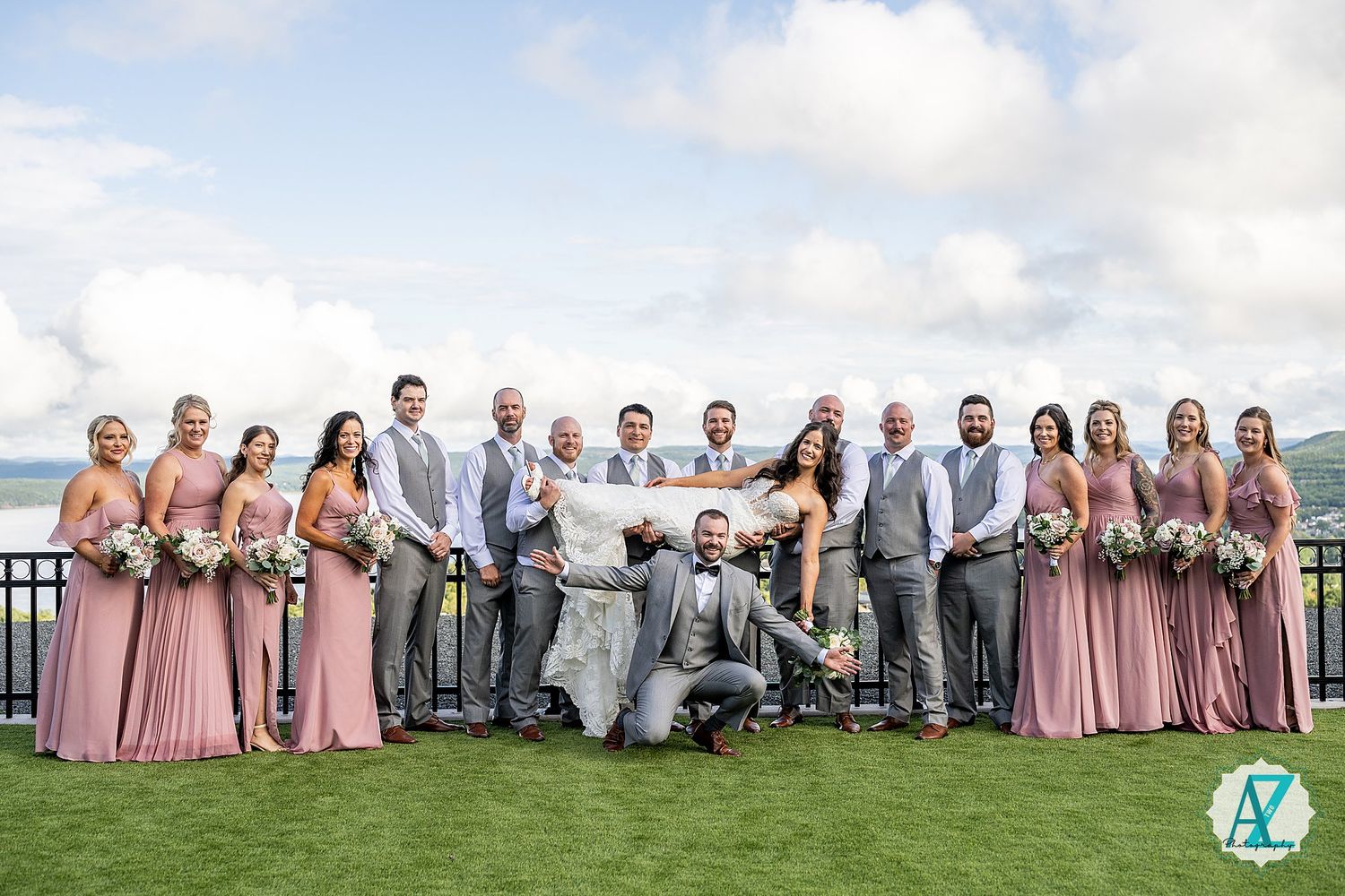 Wedding party lounges on grass for casual group photo with bridesmaids in pink and groomsmen in grey.