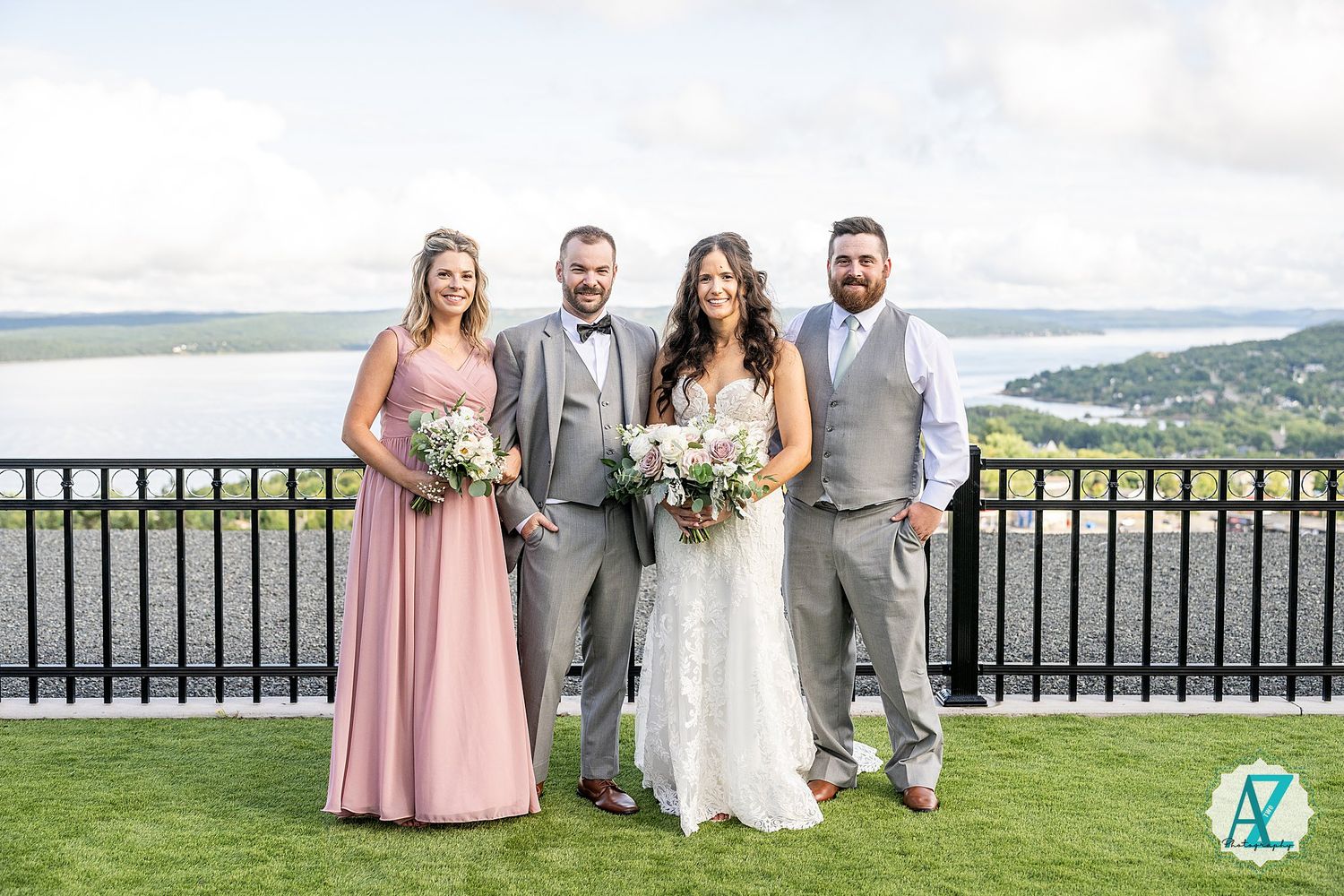 A wedding party poses together on a hilltop terrace overlooking a scenic mountain landscape on a sunny day.