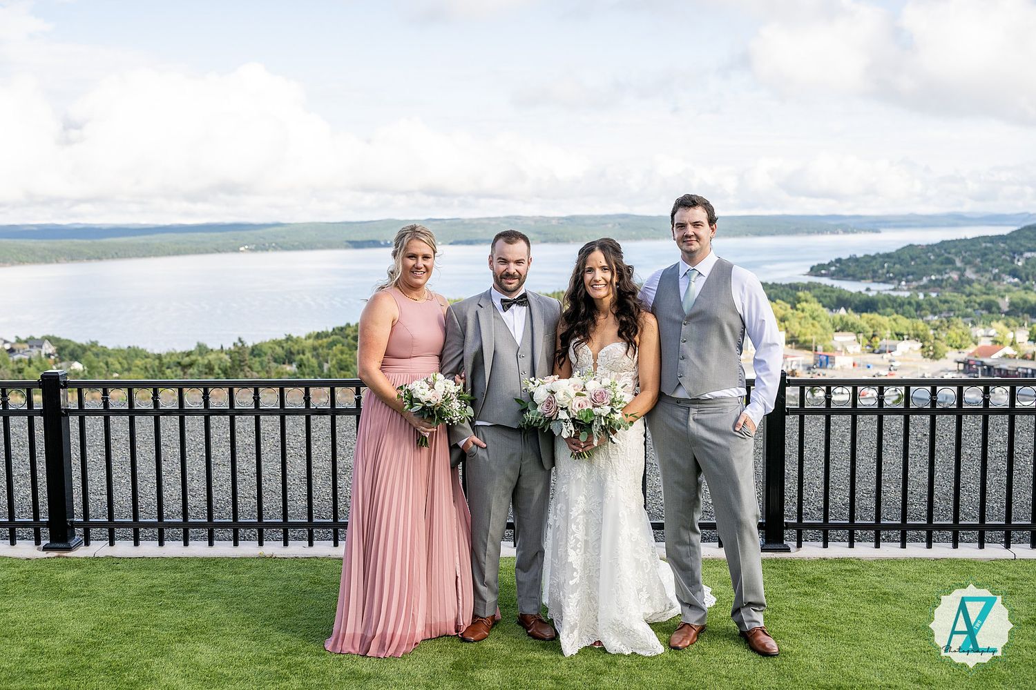 People gather on a picturesque waterfront terrace overlooking a bay with mountains and lush greenery in the background.