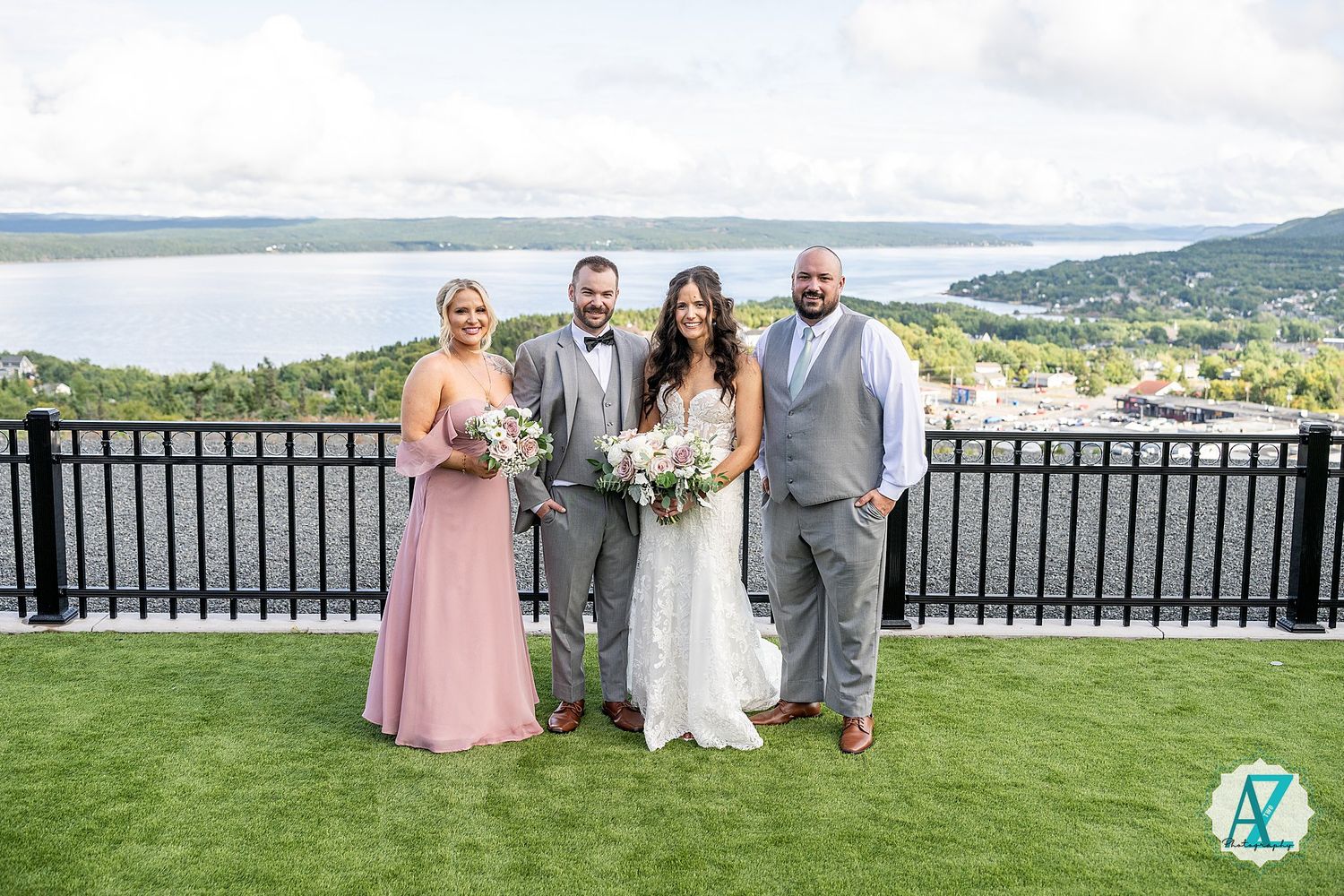 Wedding party poses on a scenic hilltop terrace overlooking water and mountains on a cloudy day.