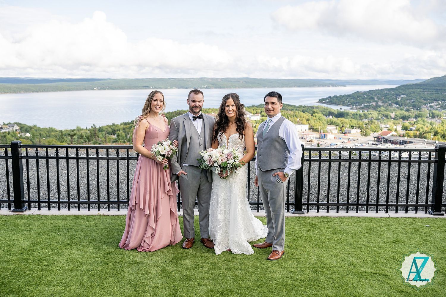 Wedding group in pink and gray attire poses outdoors with scenic mountain views behind black railing.