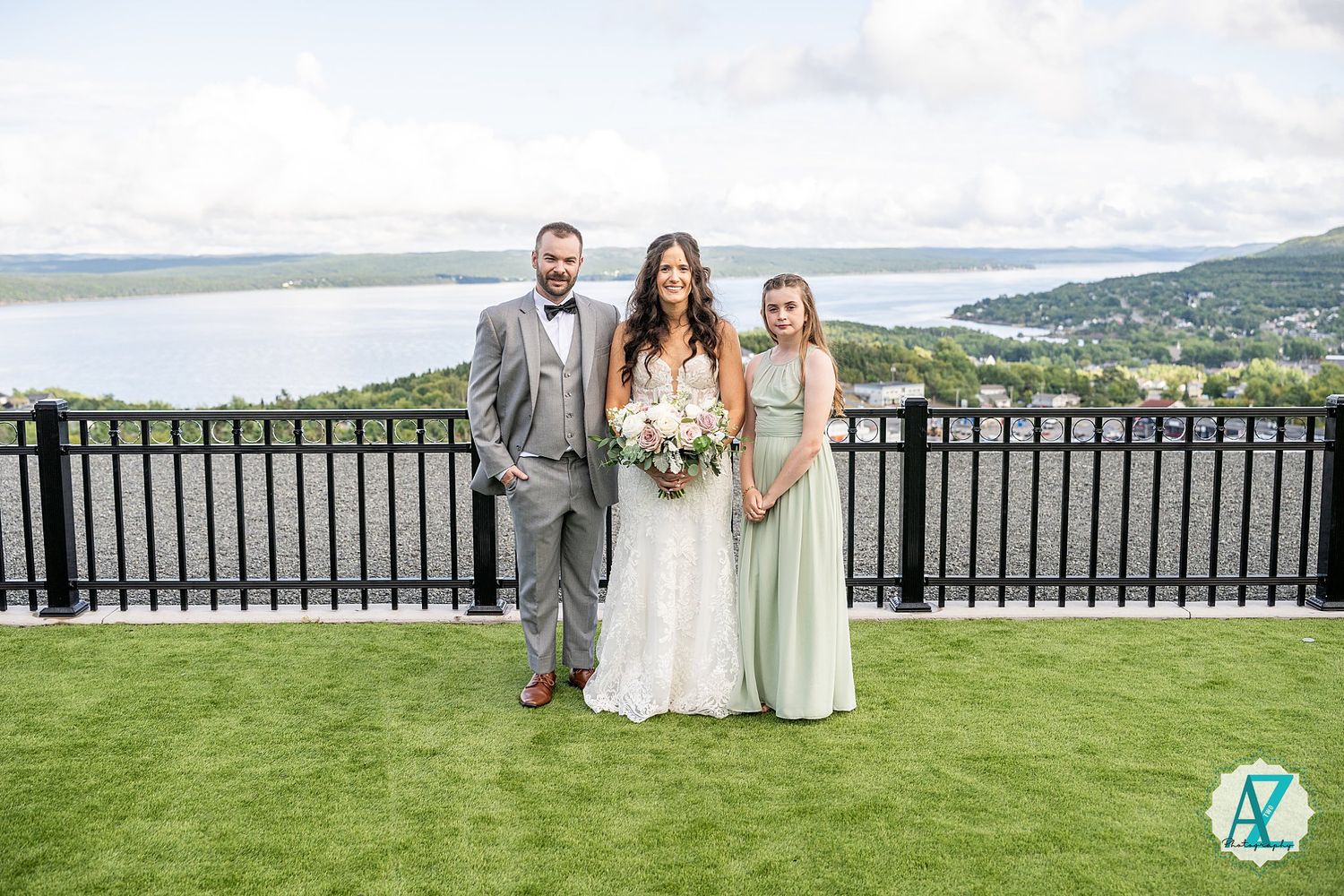 Group gathers for outdoor wedding photos on a grassy terrace with panoramic lake and mountain backdrop.