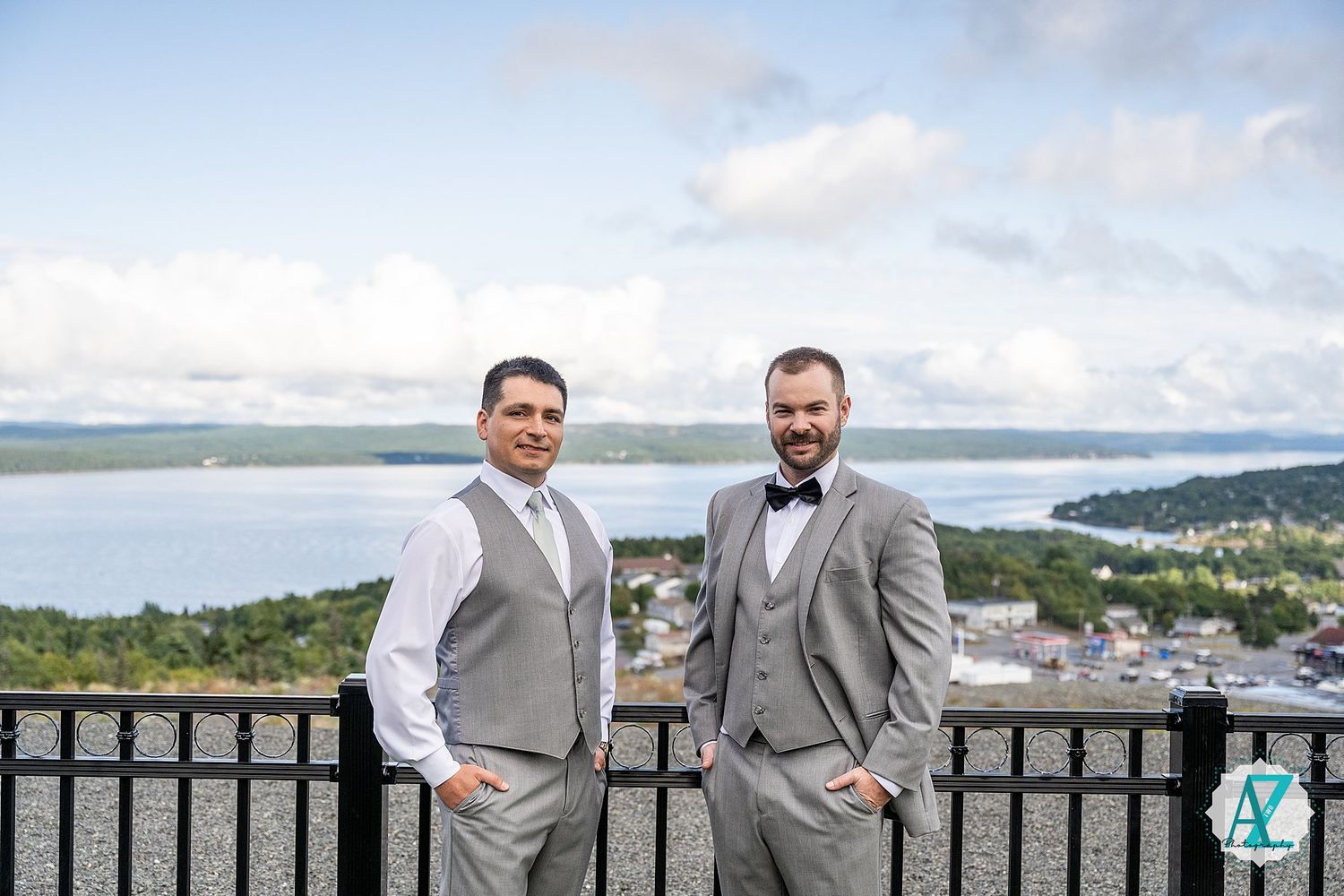 Two people in grey suits and bowties stand on an overlook with water and mountains in the background.