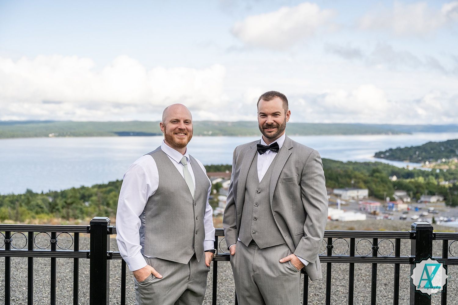 Groomsmen in grey suits and bowties posing on an outdoor terrace overlooking water.