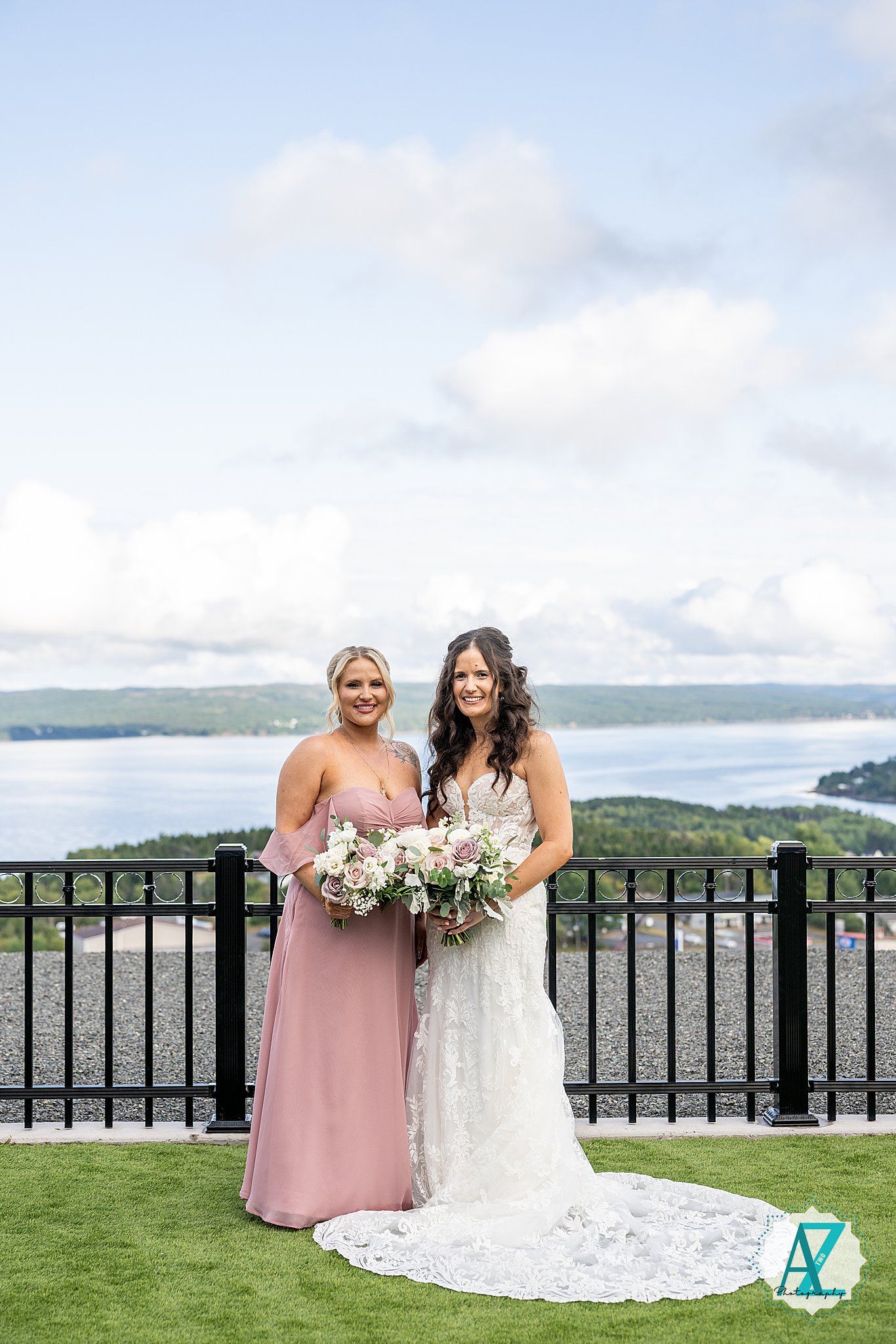 Two women in formal dresses pose together on an outdoor terrace overlooking a scenic lake and mountains.