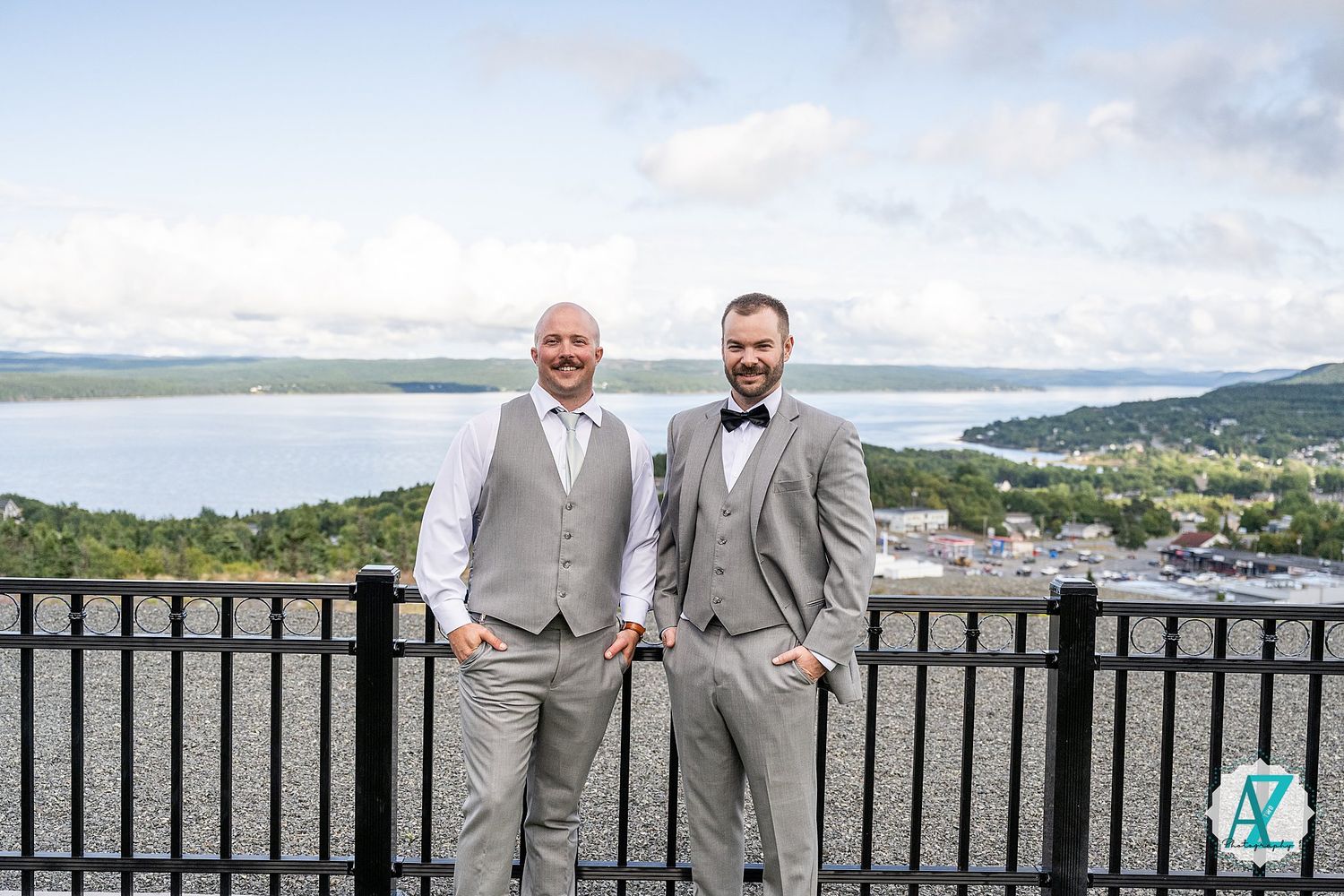 Wedding party members in light gray suits stand on a deck overlooking a scenic mountain landscape on a cloudy day.