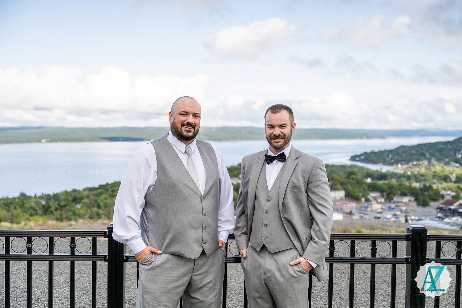 Groomsmen in light gray suits gather on a scenic overlook deck with water views in the background.