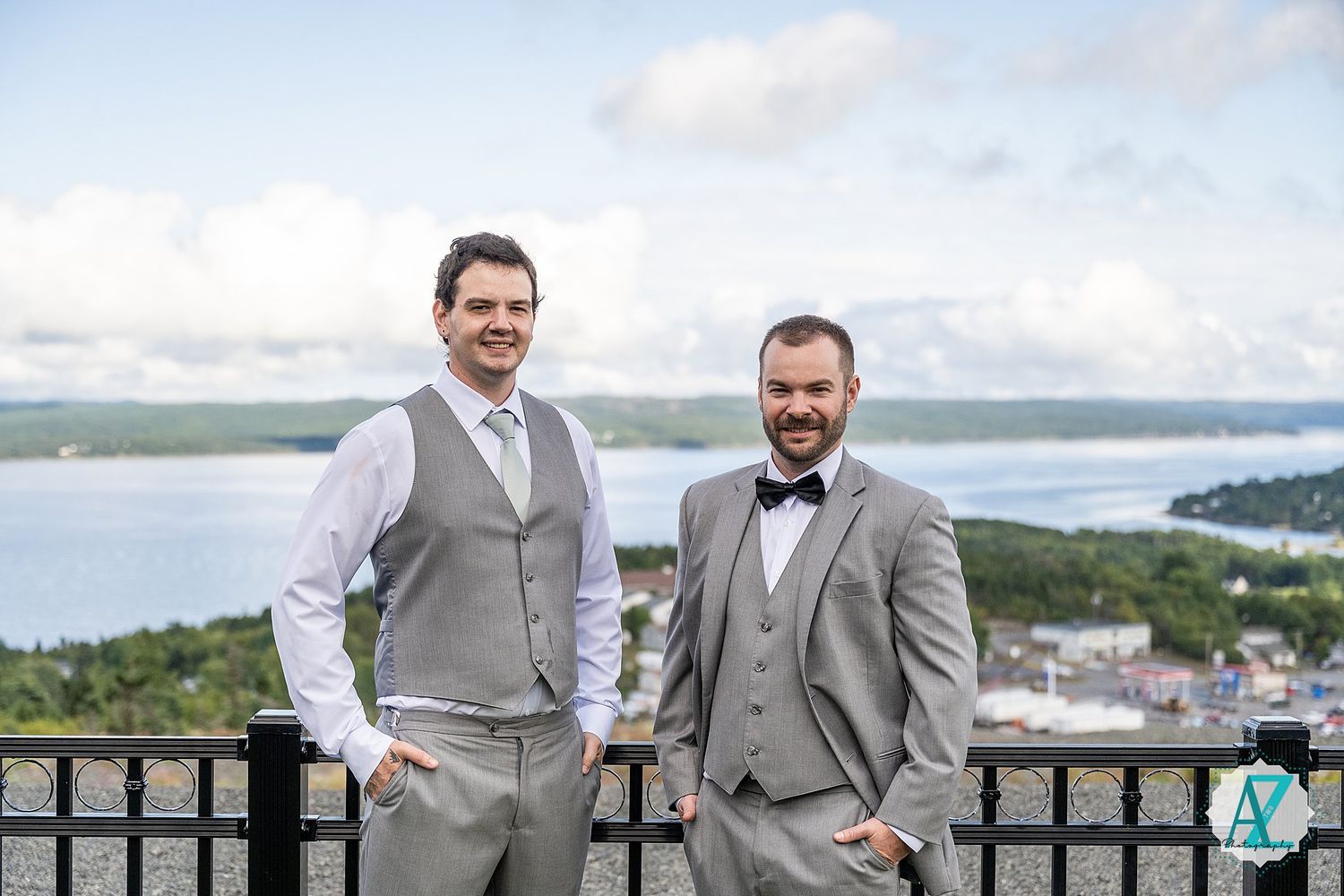 Groomsmen in light grey vests and suits standing on a scenic overlook with water views.