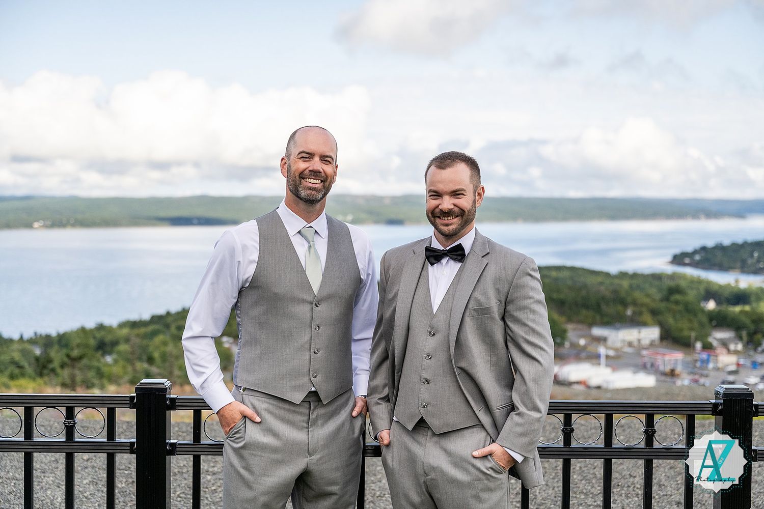 Wedding party in matching grey suits pose together on a scenic overlook with water and coastline in the background.