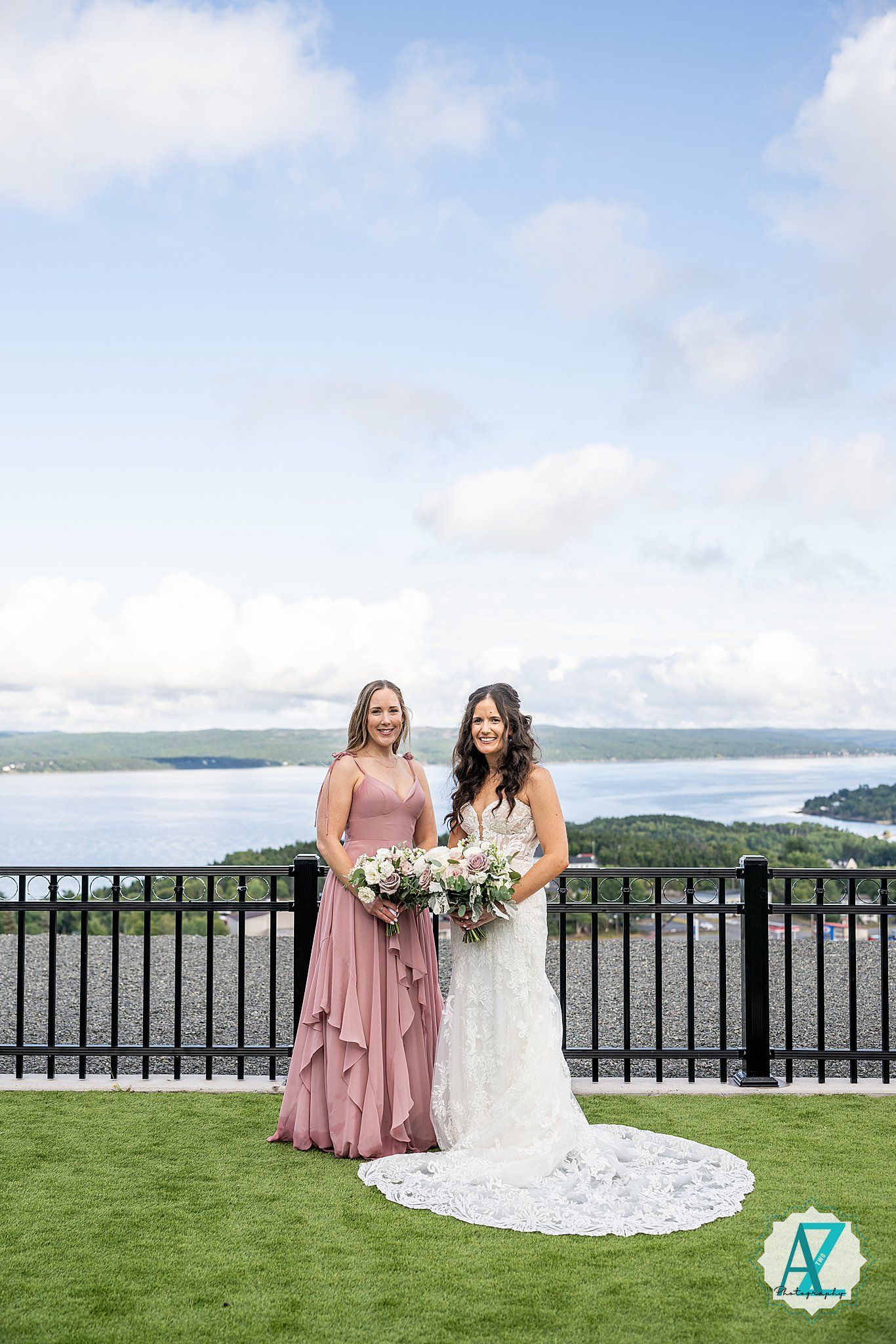 Wedding party in pink and white dresses pose against black railing with scenic water view.