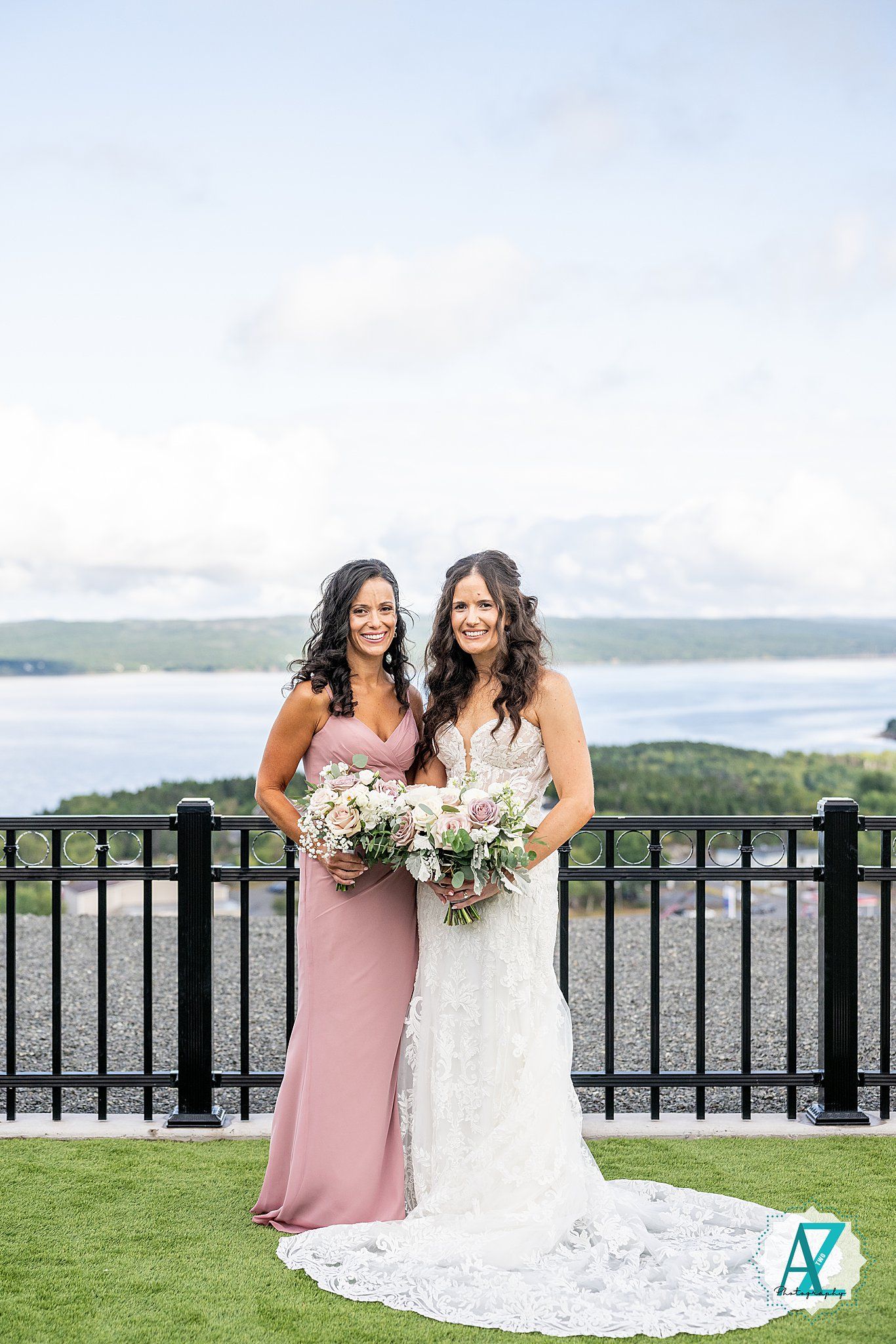 Wedding party in blush and white dresses pose on balcony overlooking scenic waterfront view.