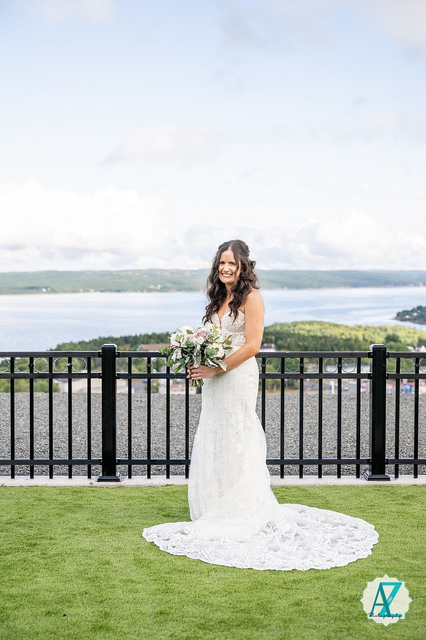 Bride in flowing lace gown poses on outdoor terrace with water view.