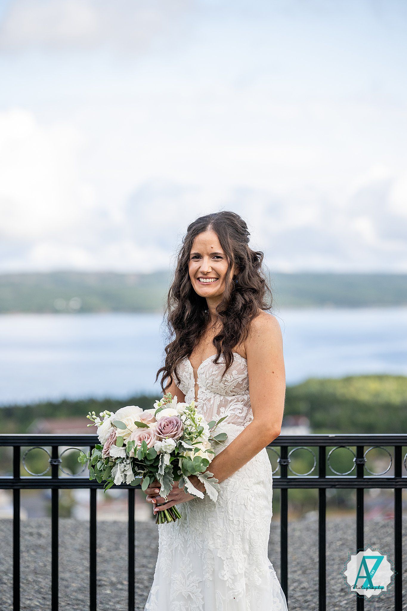 A bride in a lace wedding dress holds a purple and white bouquet while posing on an overlook with water views.