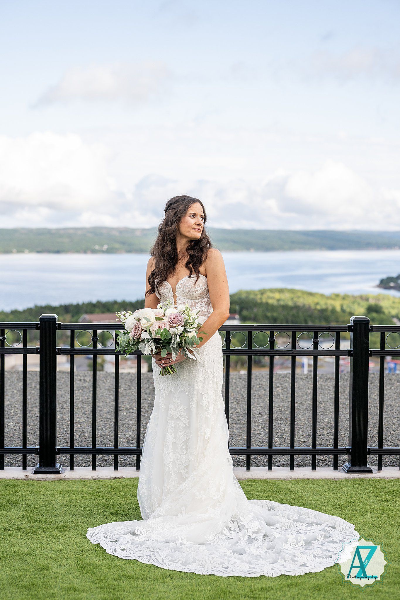 Bride in lace gown with flowing train poses against scenic overlook with water views.