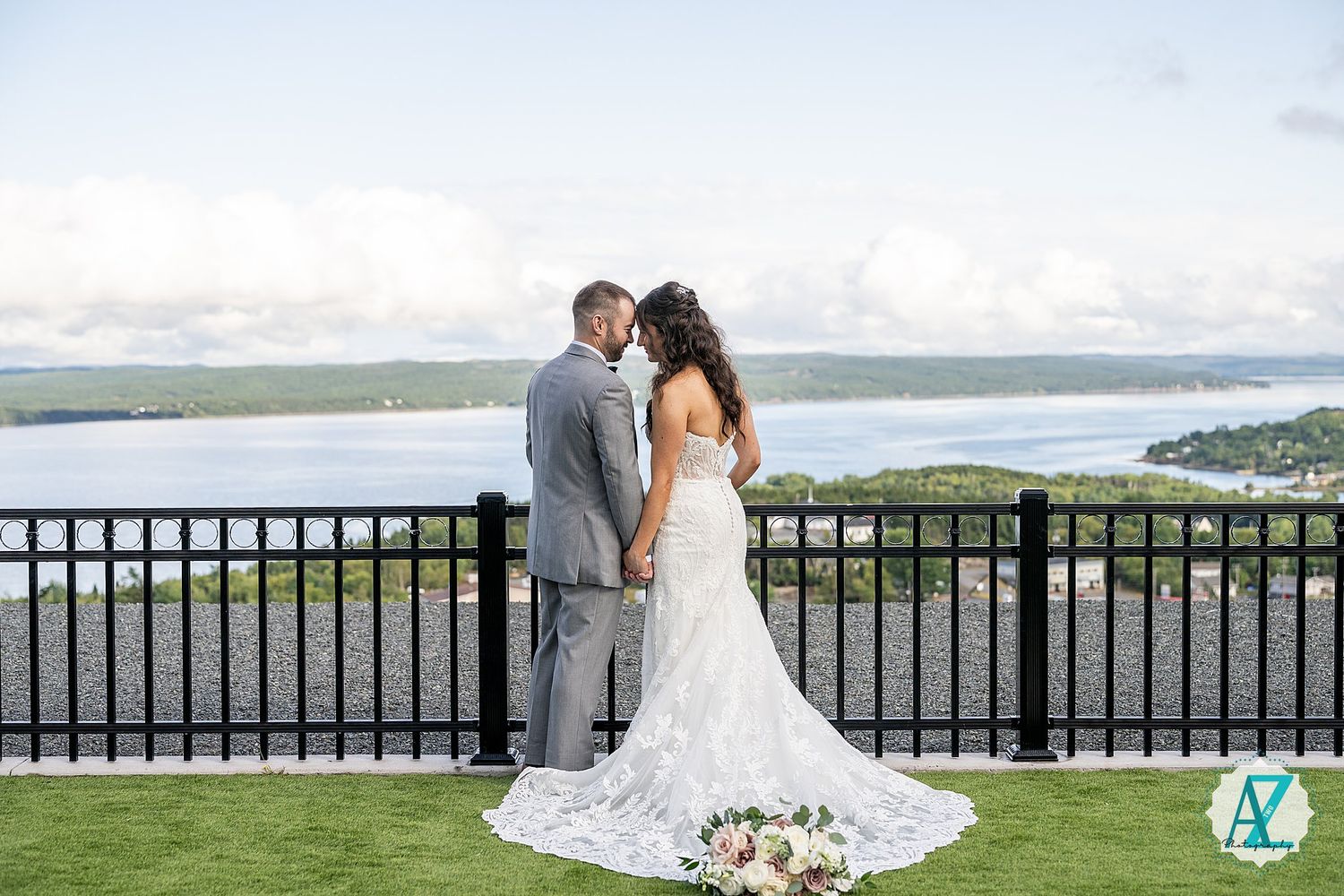 People gather for wedding photos on a grassy overlook with wrought iron railing and stunning water views.