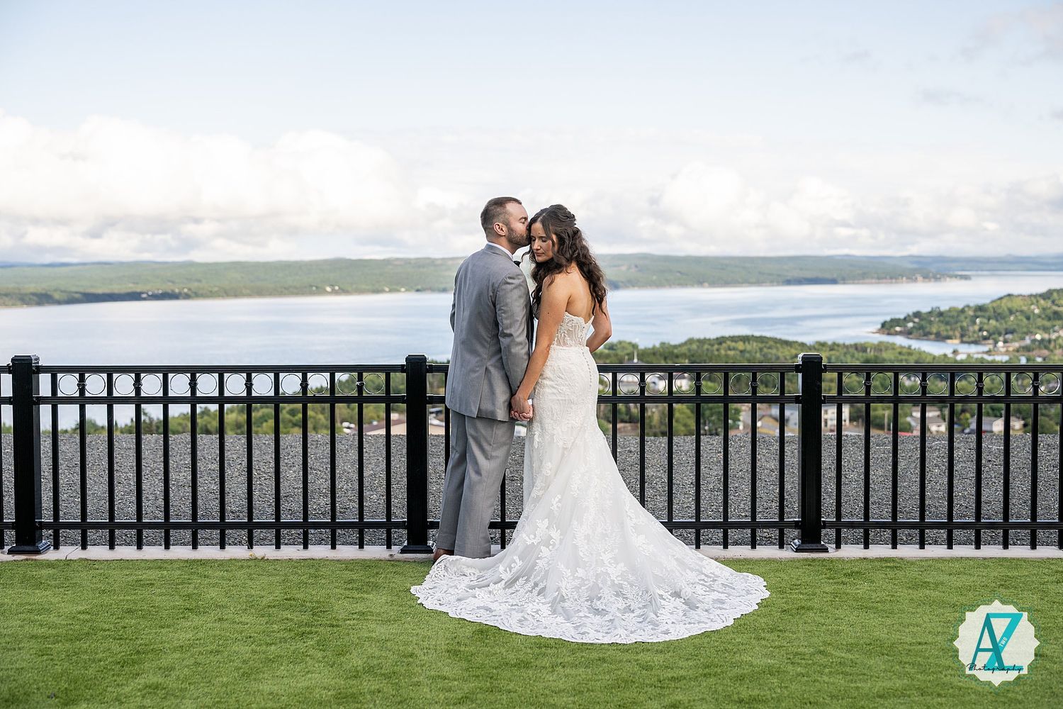 A couple stands on a scenic overlook with water views and lush green grass against a railing on their wedding day.