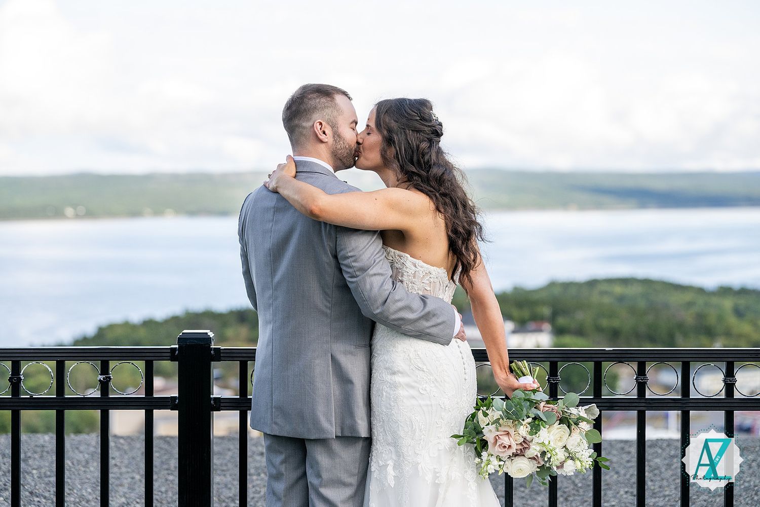Couple in wedding attire embraces on scenic overlook with lake view and bouquet in hand.