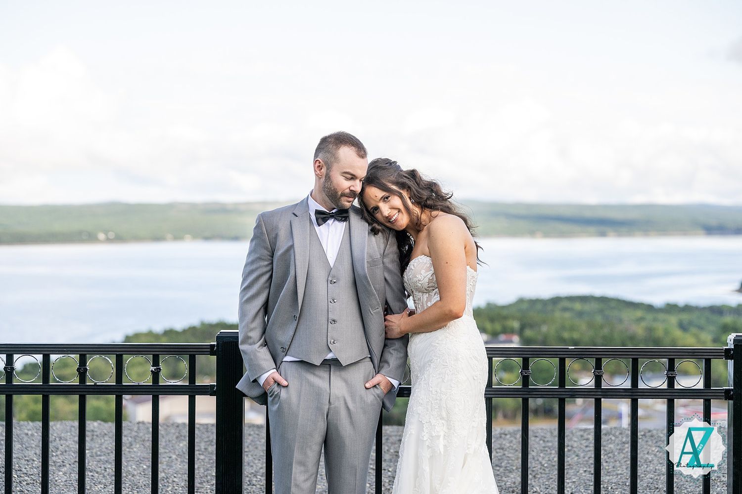 A romantic couple embraces on a stone balcony overlooking a scenic waterfront view on their wedding day.