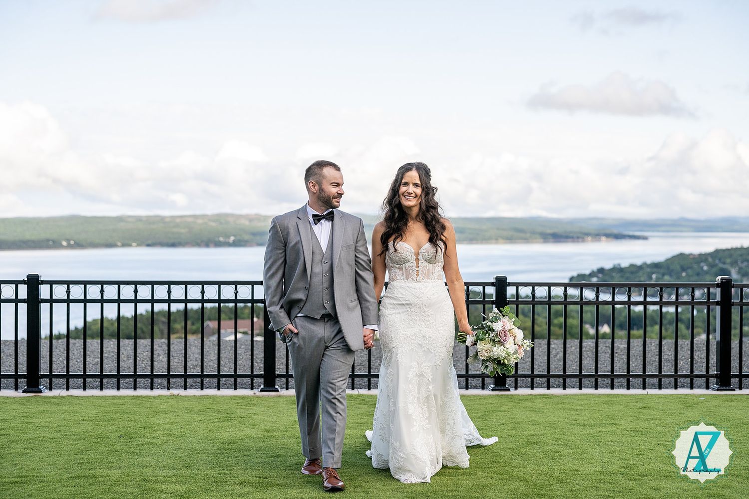 A couple shares a romantic moment along a waterfront railing with scenic views of rolling hills in the background.