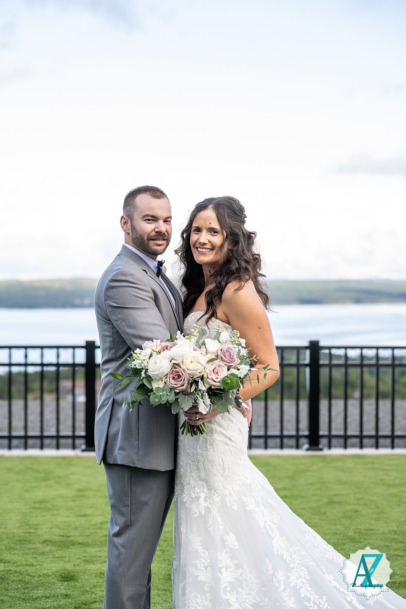 A couple in wedding attire share joyful laughter outdoors with a scenic waterfront view in the background.