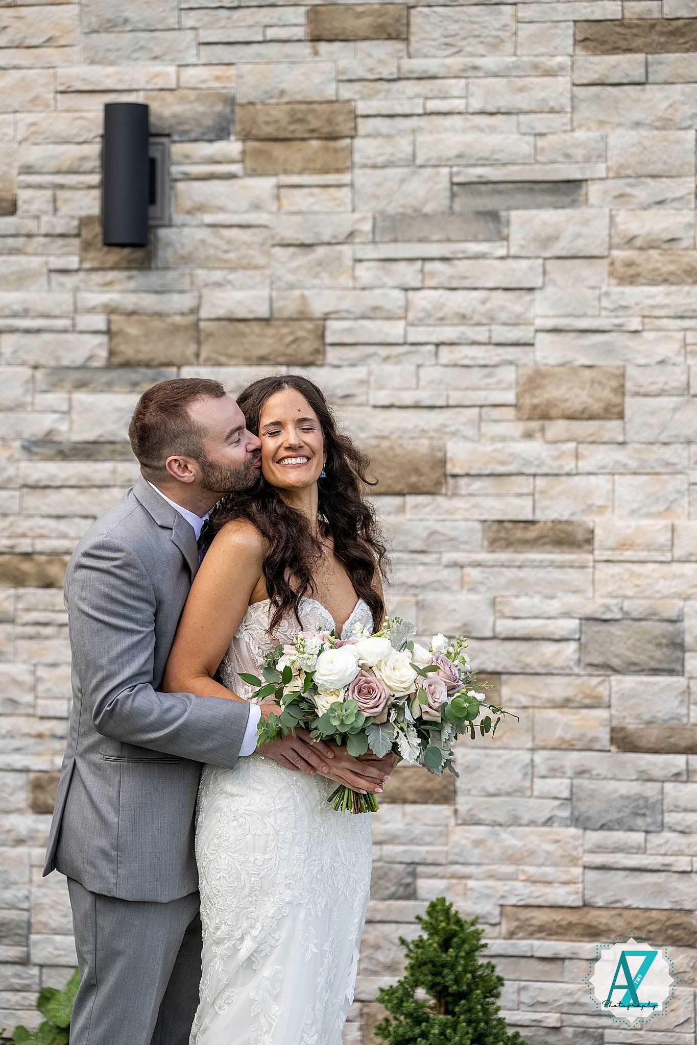 A wedding couple shares romantic moments against a stone wall while holding a pastel floral bouquet.