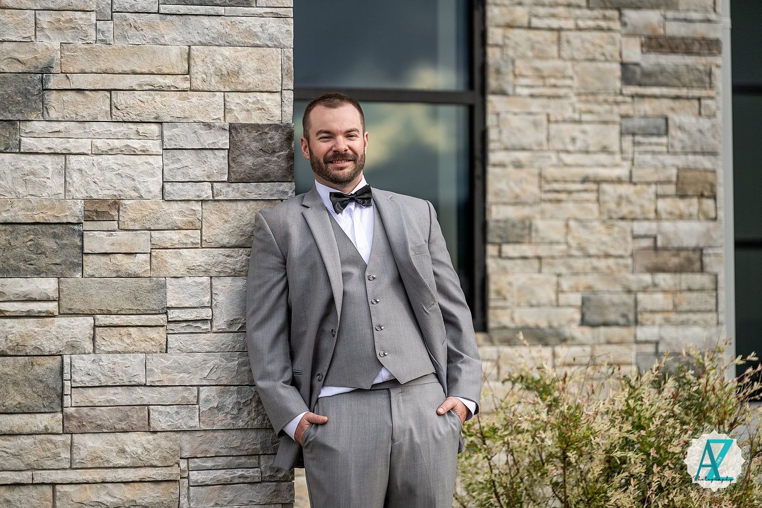 A groom in a grey three-piece suit stands confidently outside a stone building during a wedding photoshoot.