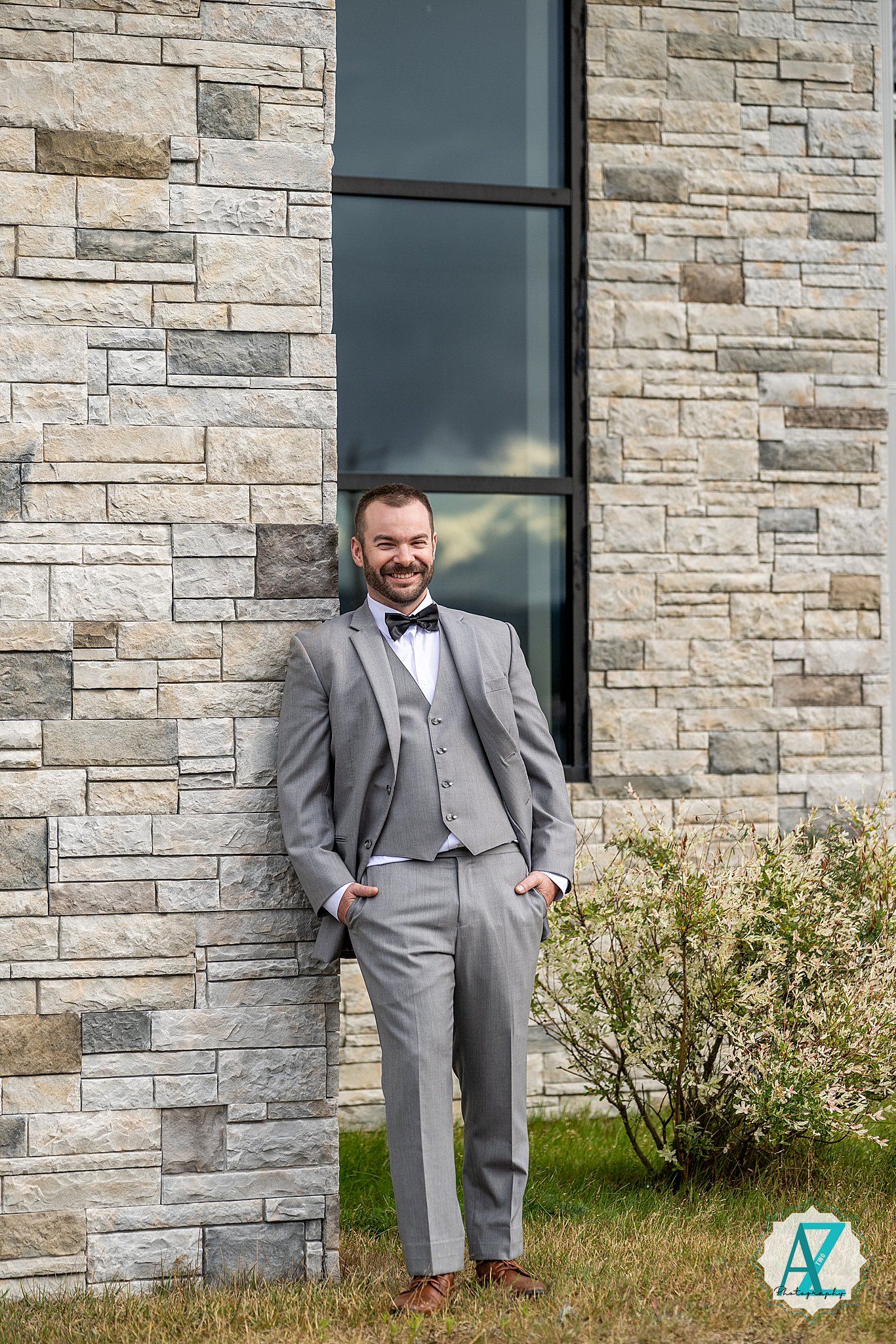 A person in a gray suit and bow tie poses confidently against a stone wall exterior of a building.