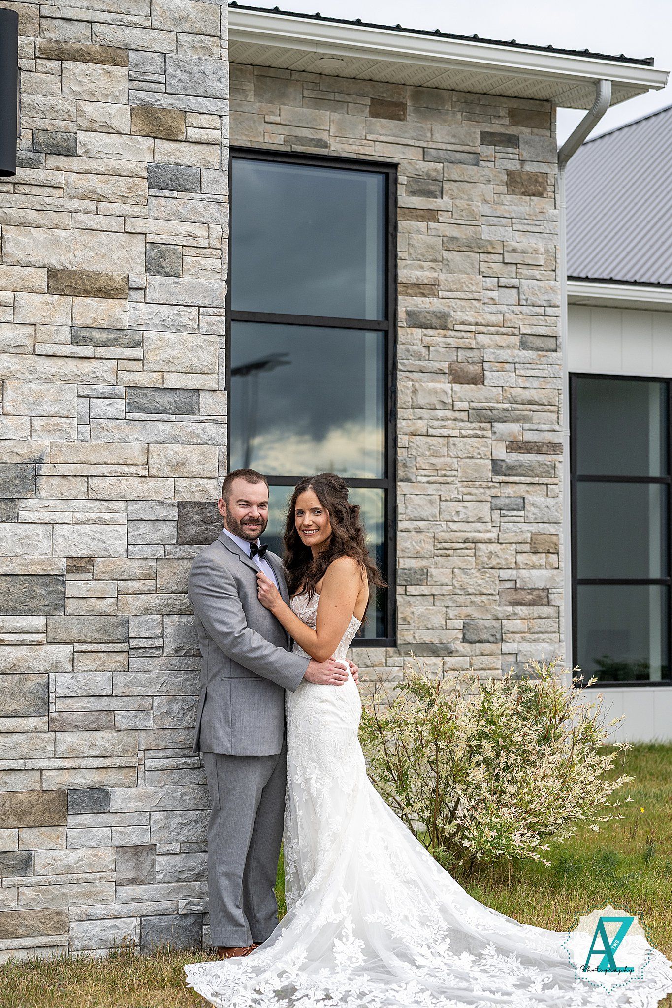 A couple poses together against a modern stone wall exterior of a house with large windows.