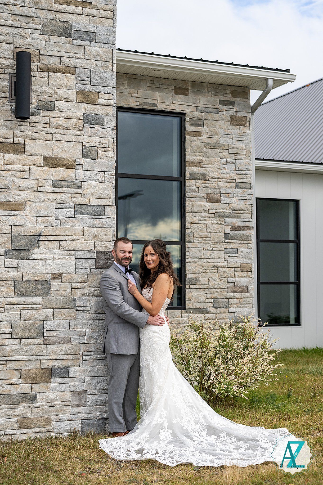 A wedding photo featuring a couple posing against a modern stone exterior wall with a long flowing white lace dress.