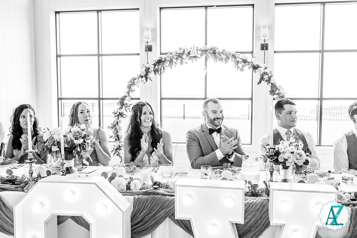 Wedding party seated at long white table during reception with floral decorations and bright windows in background.