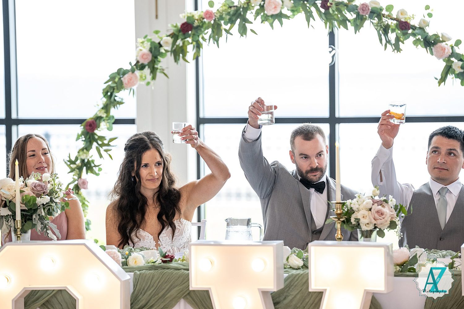 Wedding celebration scene in a bright venue with floral garlands and people raising glasses.