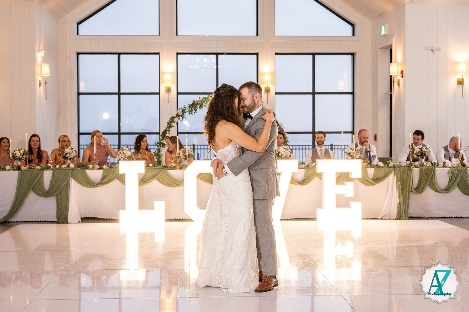 Silhouetted couple in an embrace dance together in an elegant white venue with a bridal party looking on.