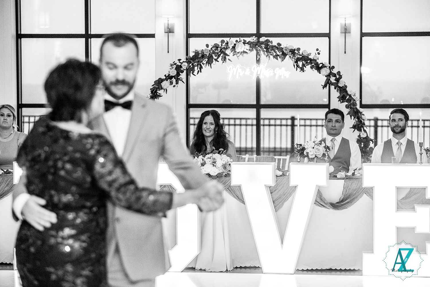 Black and white photo of a wedding celebration with floral arch and LOVE sign visible in background.