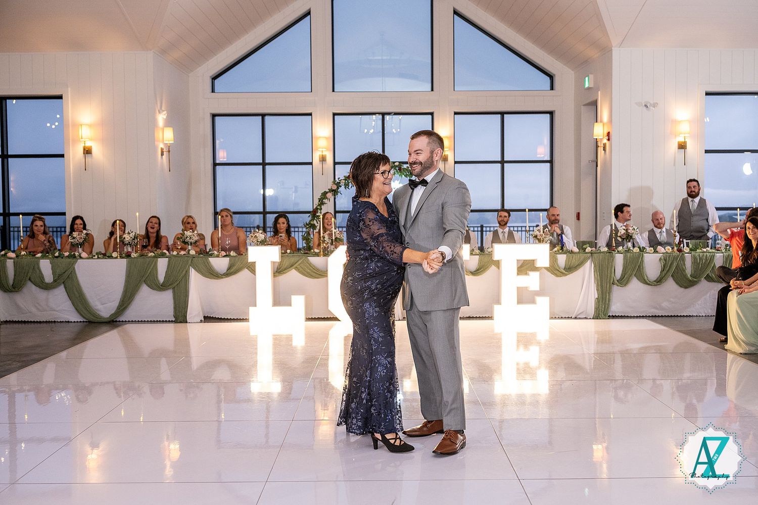 People dance together during a celebration in a bright, airy room with large windows and pendant lighting.