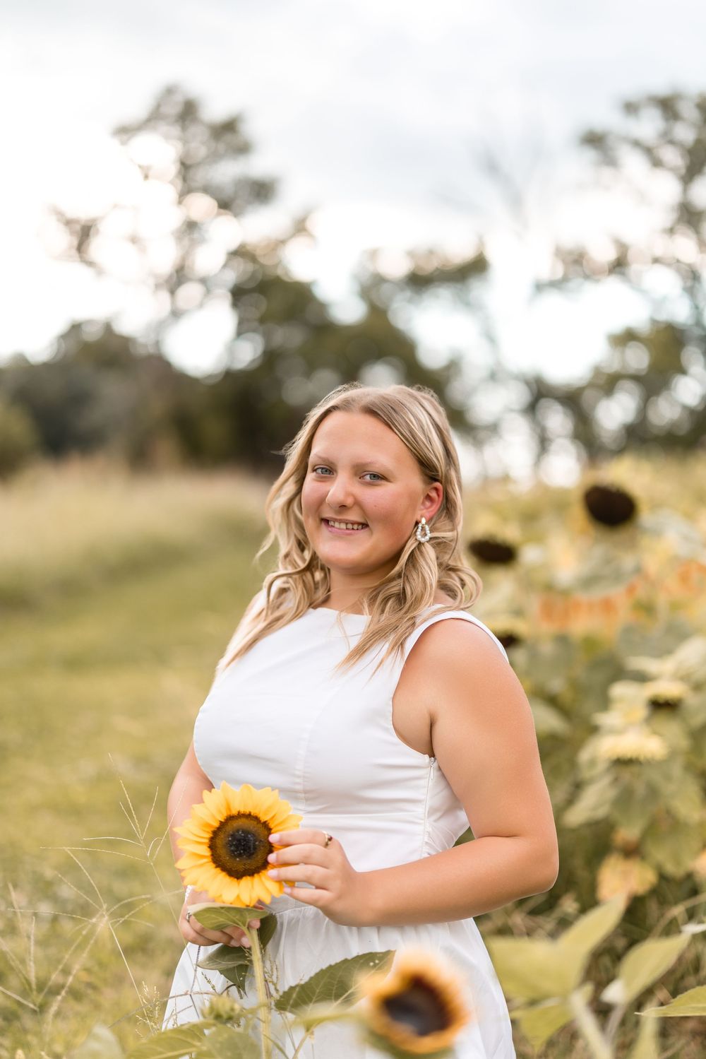 A person in a white dress stands among blooming sunflowers in a sunny outdoor field.