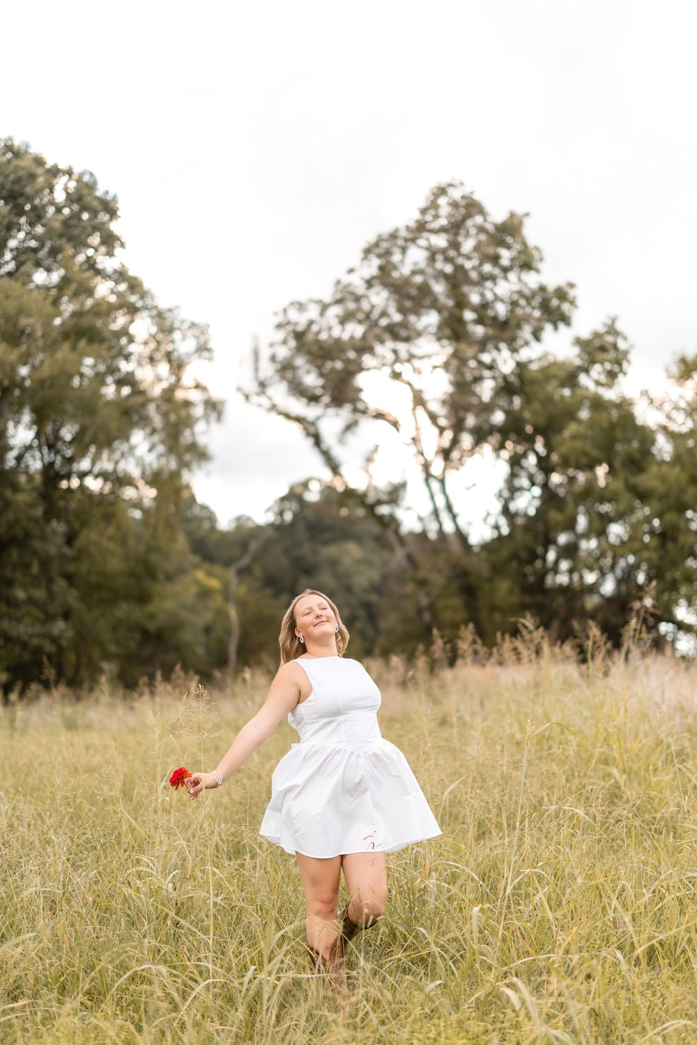 A figure in a white dress dances and twirls joyfully through a sunlit meadow with tall grass and trees in the background.