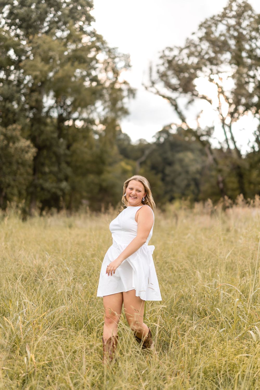Person wearing white dress spins joyfully in tall grass field with trees silhouetted against sky.
