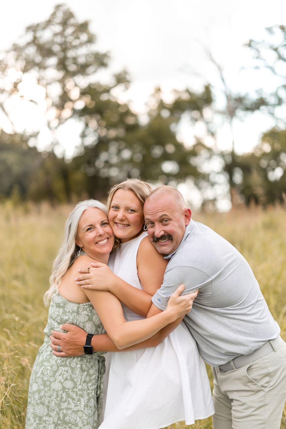 Three people sharing a joyful embrace and laughing together in a sunny field with tall grass.