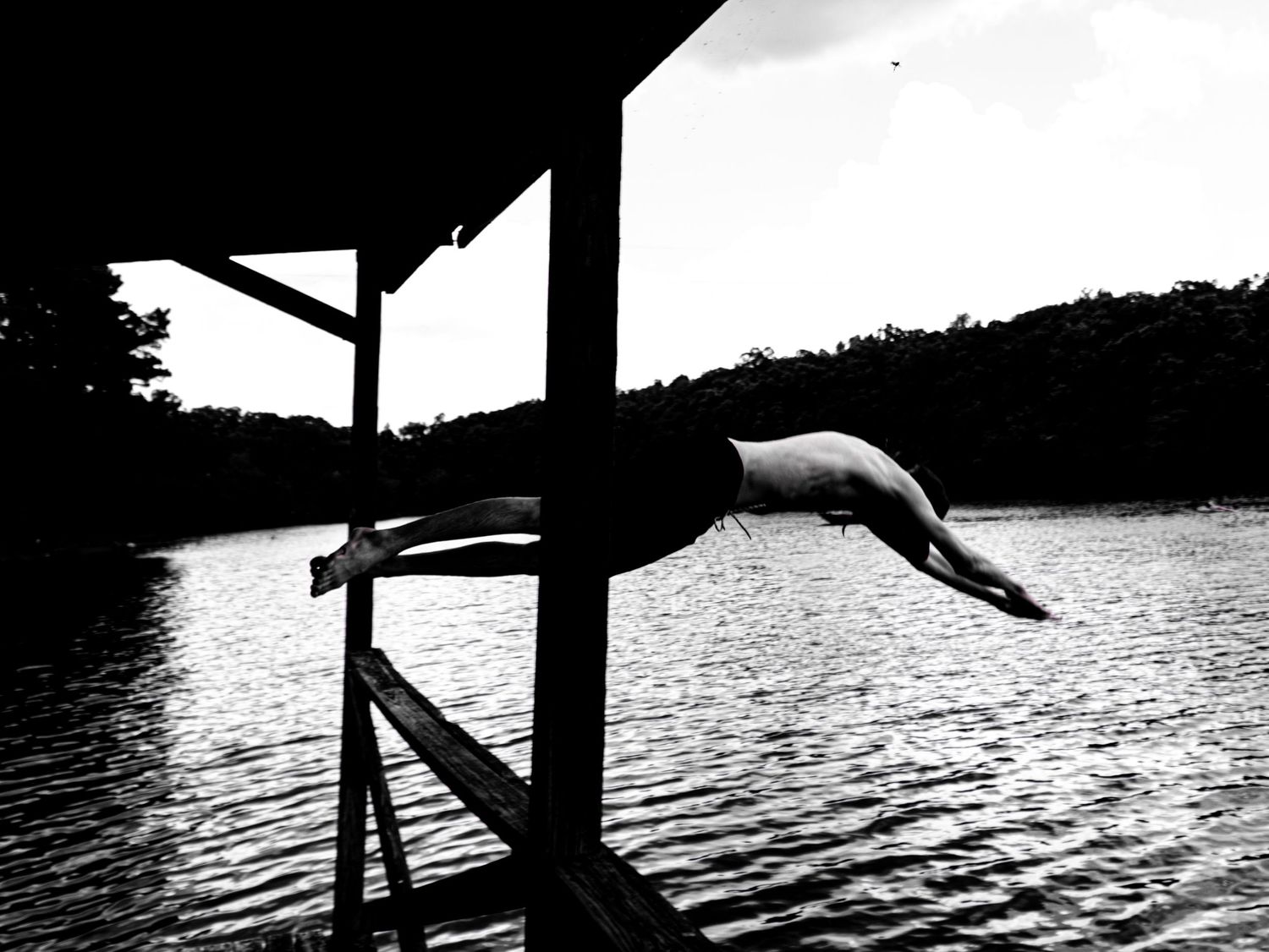 A black and white panoramic silhouette of a wooden pier extending over a tranquil lake with trees lining the horizon.