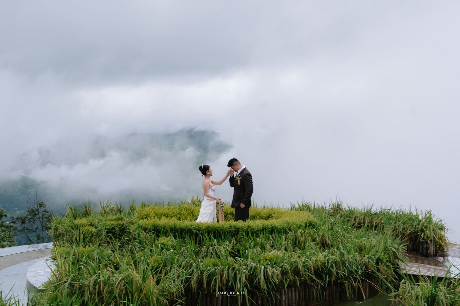 Wedding couple sharing a joyful moment on elevated green terrace surrounded by dramatic misty mountain clouds.